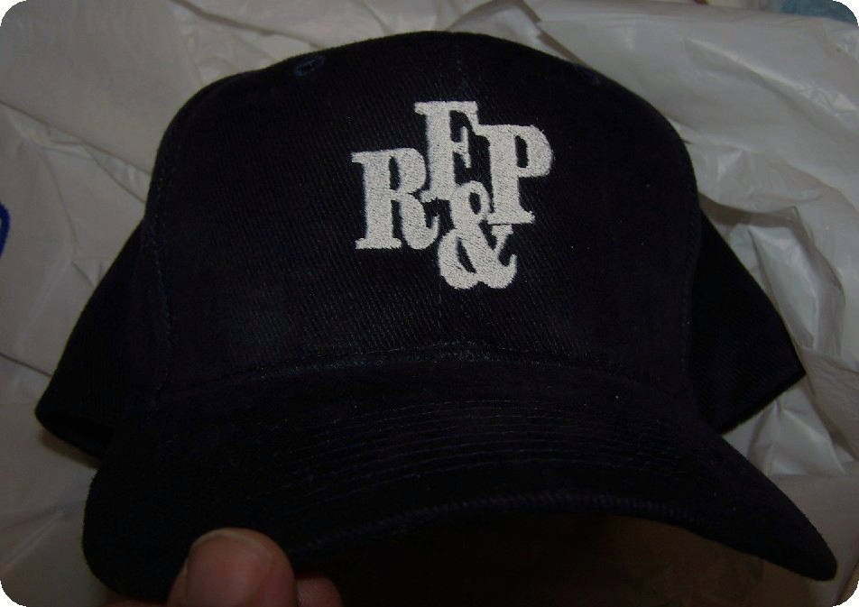 Photo of a baseball cap in navy, with white RF&P logo from the 1980s on front held by human hand above a white background.