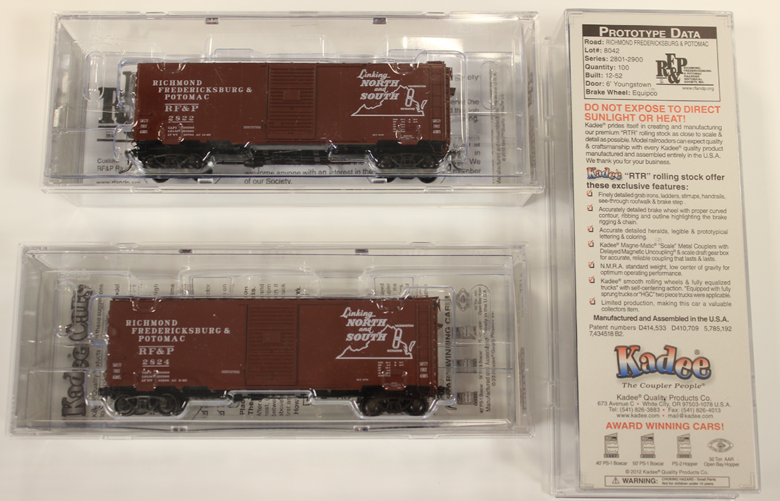Photo of two HO scale boxcars in red-brown with the RF&P map herald logo. Two boxcars are in clear plastic shipping boxes. A third shipping box sits alongside, showing the bottom of the box with text describing the cooperation between Kadee and the RF&PRRHS.